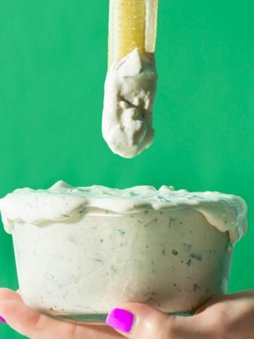 A hand with bright purple nail polish holds a glass bowl overflowing with creamy dip, as a celery stick coated in dip is lifted above the bowl against a vibrant green background.