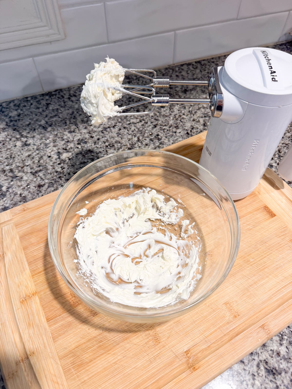 A hand mixer with beaters coated in cream cheese rests above a glass bowl containing partially mixed cream cheese, all placed on a wooden cutting board on a granite countertop.