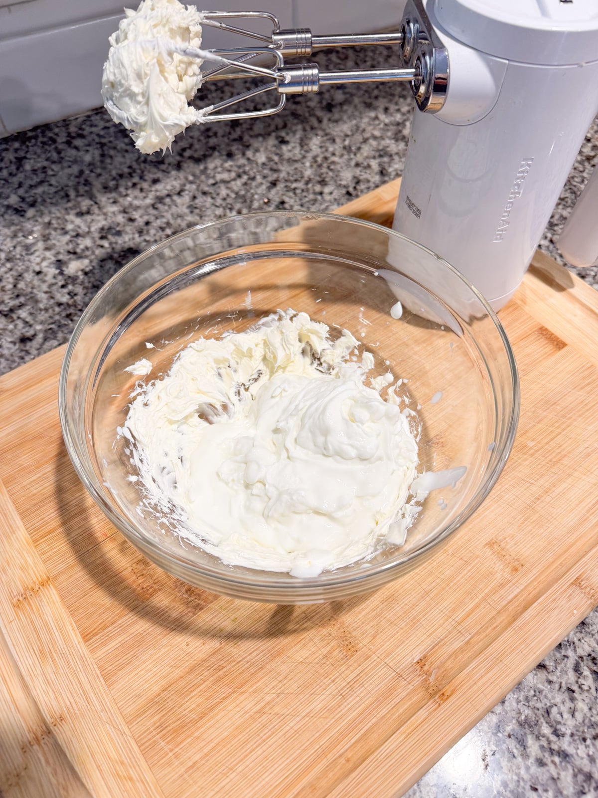 A glass bowl with whipped cream sits on a wooden cutting board. An electric hand mixer with beaters covered in whipped cream rests nearby on a kitchen counter.