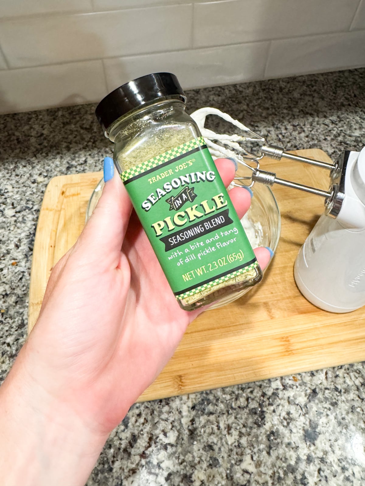 A hand holds a bottle of Trader Joe's Seasoning in a Pickle blend over a wooden cutting board, with a glass bowl and white hand mixer on a granite countertop in the background.