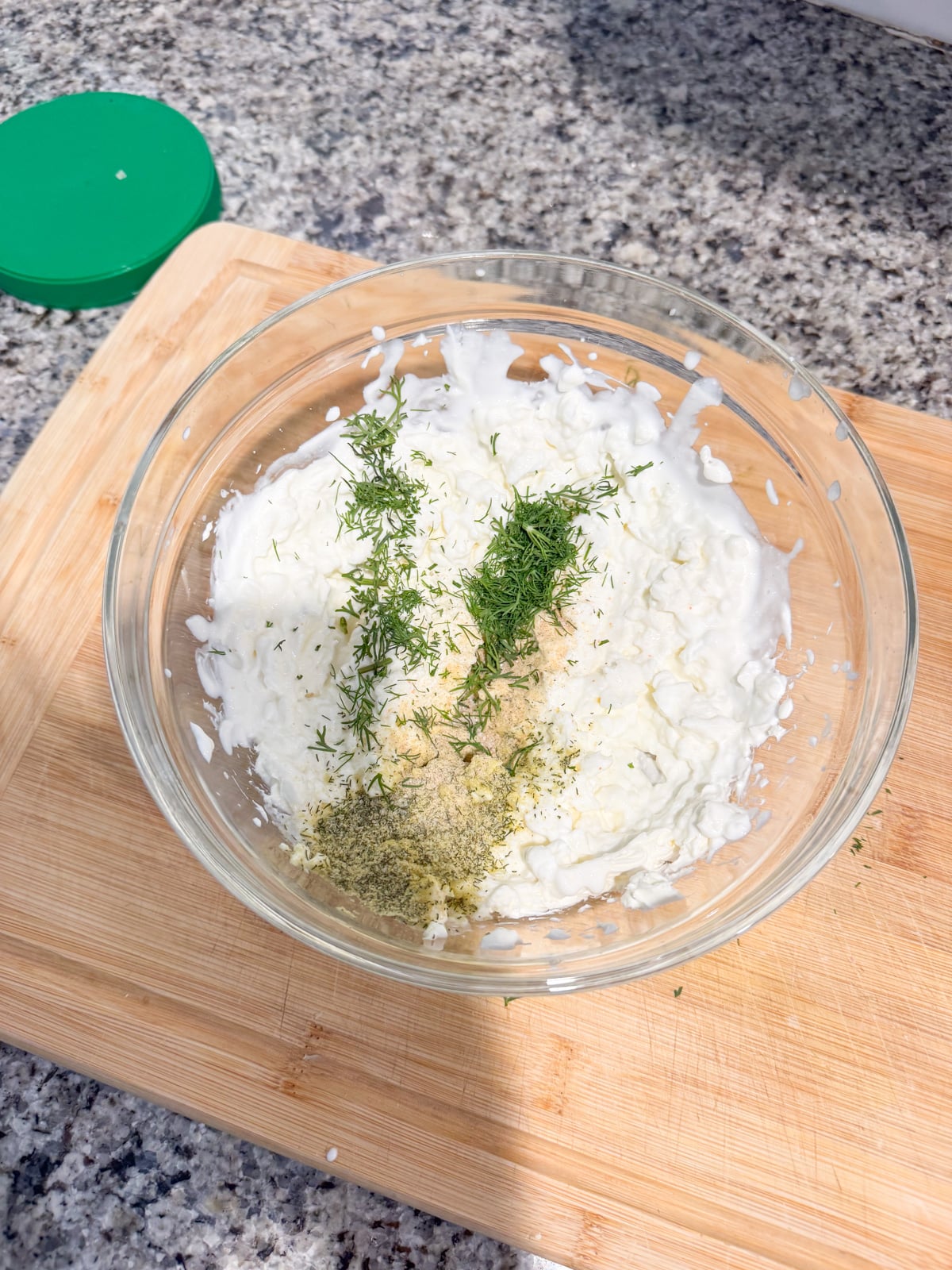 A glass bowl on a wooden cutting board contains cottage cheese, chopped dill, black pepper, and a light-colored seasoning, ready to be mixed. The scene is set on a granite countertop with a green lid nearby.