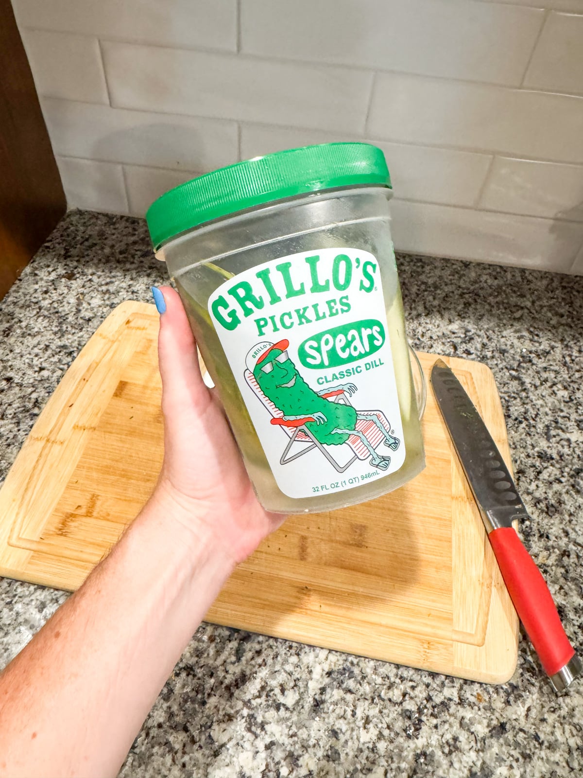 A hand holds a container of Grillo's Pickles Classic Dill Spears over a wooden cutting board and a knife with a red handle on a granite countertop.