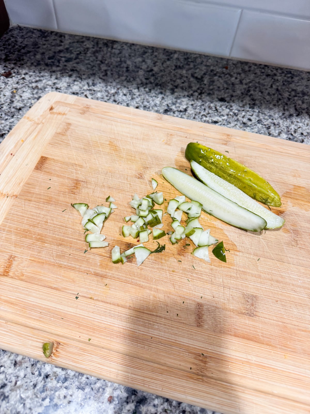 A wooden cutting board on a granite countertop with one pickle cut into spears and another pickle chopped into small pieces. White tiled backsplash in the background.