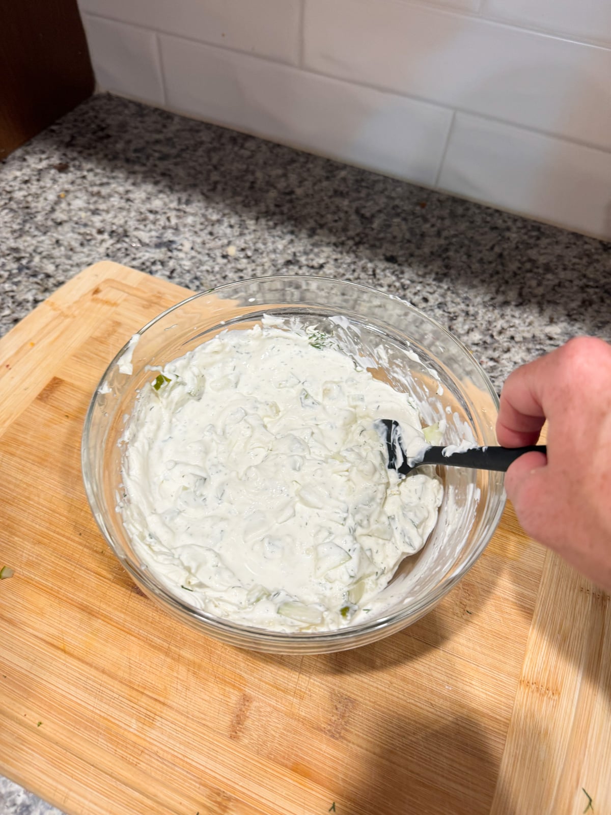 A hand stirs a bowl of creamy, white dip with a spoon on a wooden cutting board, on a speckled kitchen counter with white tiled backsplash in the background.