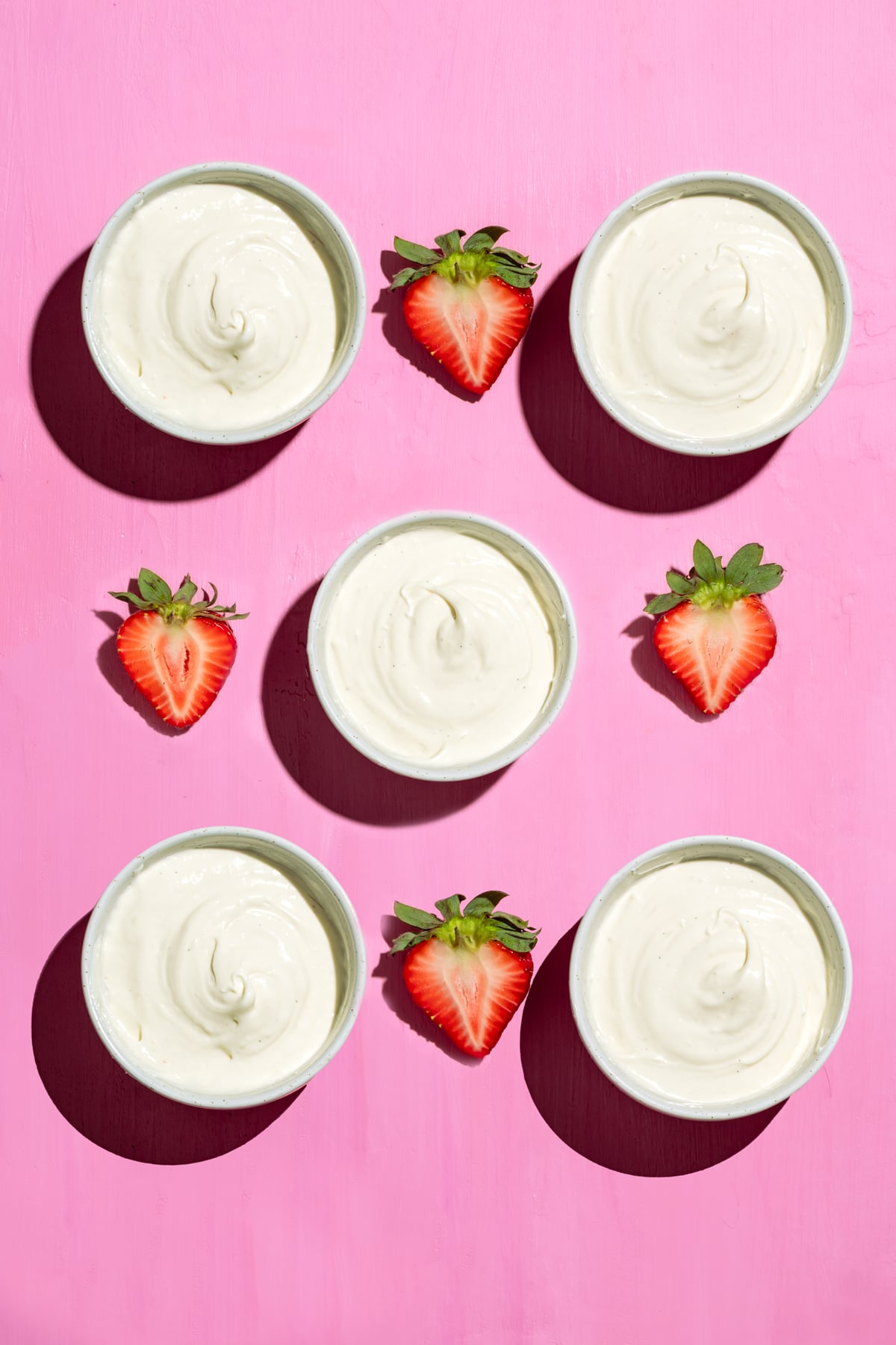 Six bowls of creamy yogurt are arranged in two rows on a pink surface, each row separated by three halved strawberries placed between the bowls.