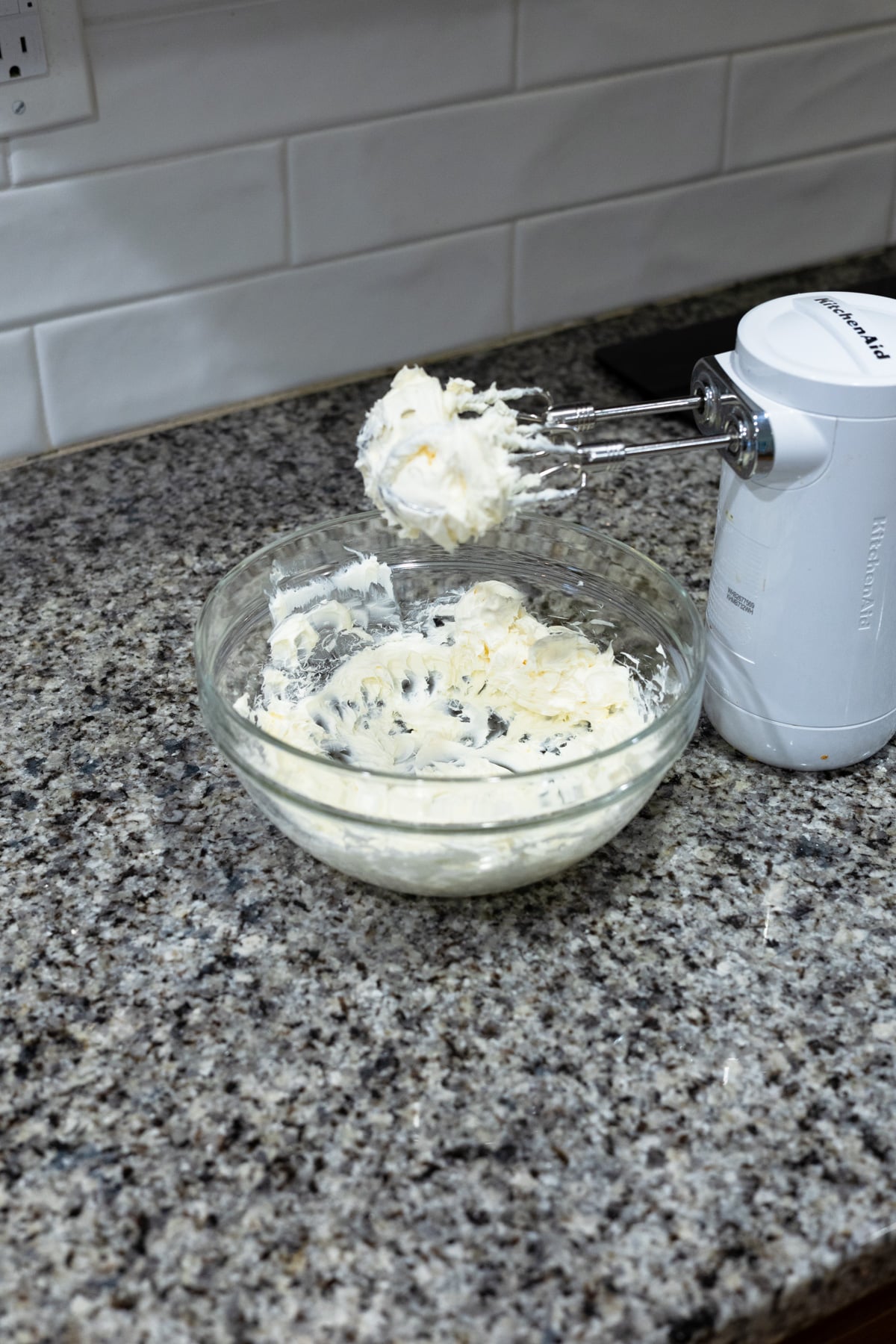 A glass bowl of whipped cream or batter sits on a granite countertop next to an electric hand mixer with beaters covered in the mixture, against a white tiled backsplash.
