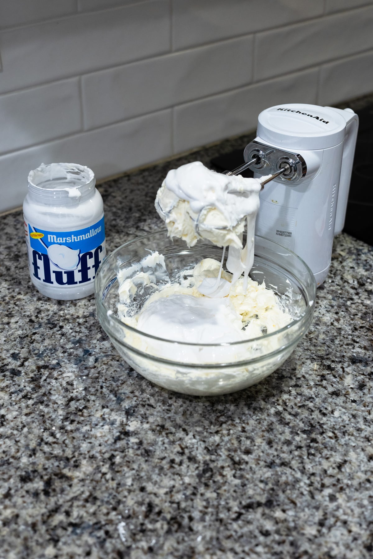 A mixing bowl with a white creamy mixture sits on a granite countertop next to an electric hand mixer and an open jar of Marshmallow Fluff. The mixer beaters are coated with the mixture.