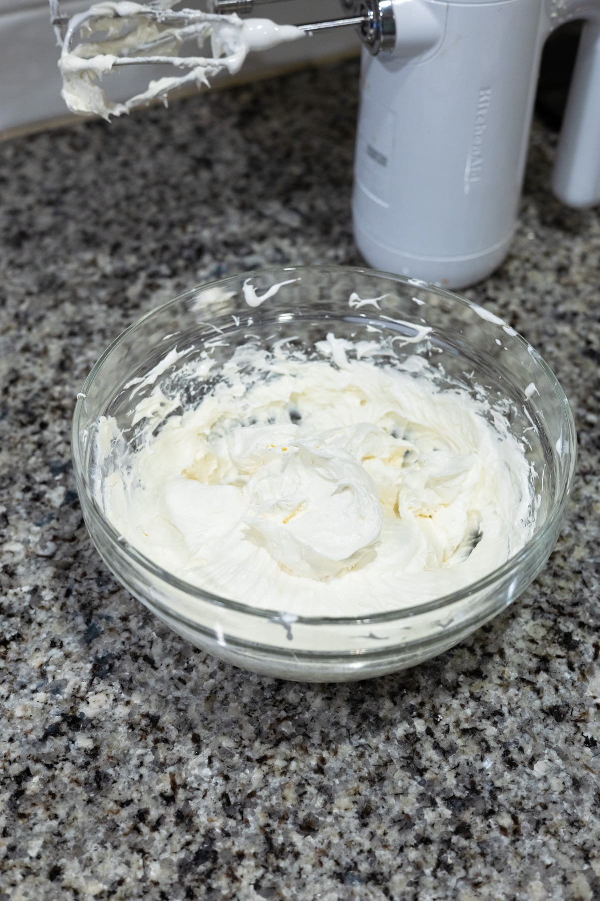 A glass bowl filled with whipped cream cheese mixture sits on a granite countertop, with a hand mixer and beaters covered in the mixture nearby.