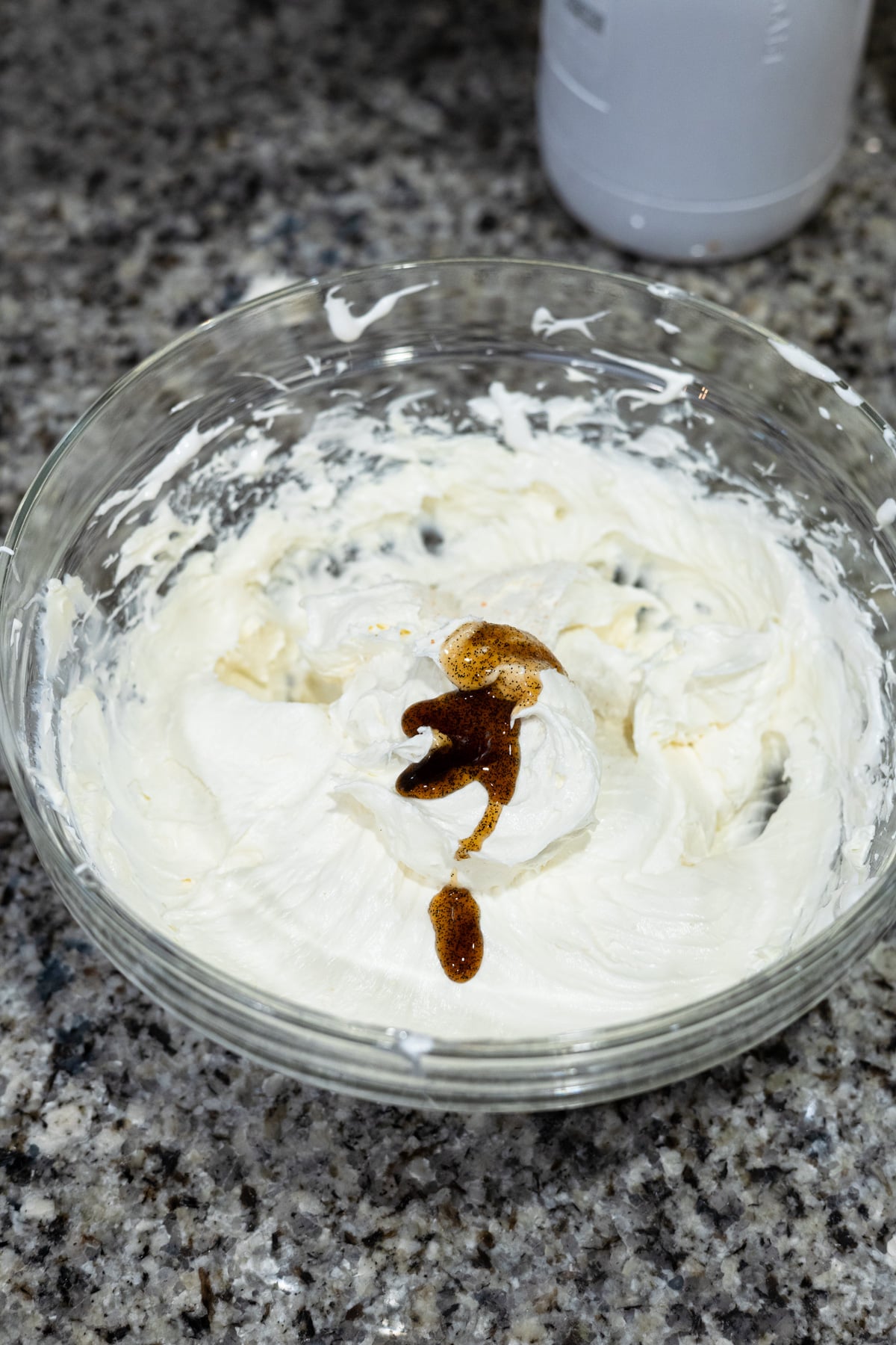 Glass bowl with whipped cream cheese mixture and a dollop of brown vanilla extract on top, sitting on a speckled granite countertop.