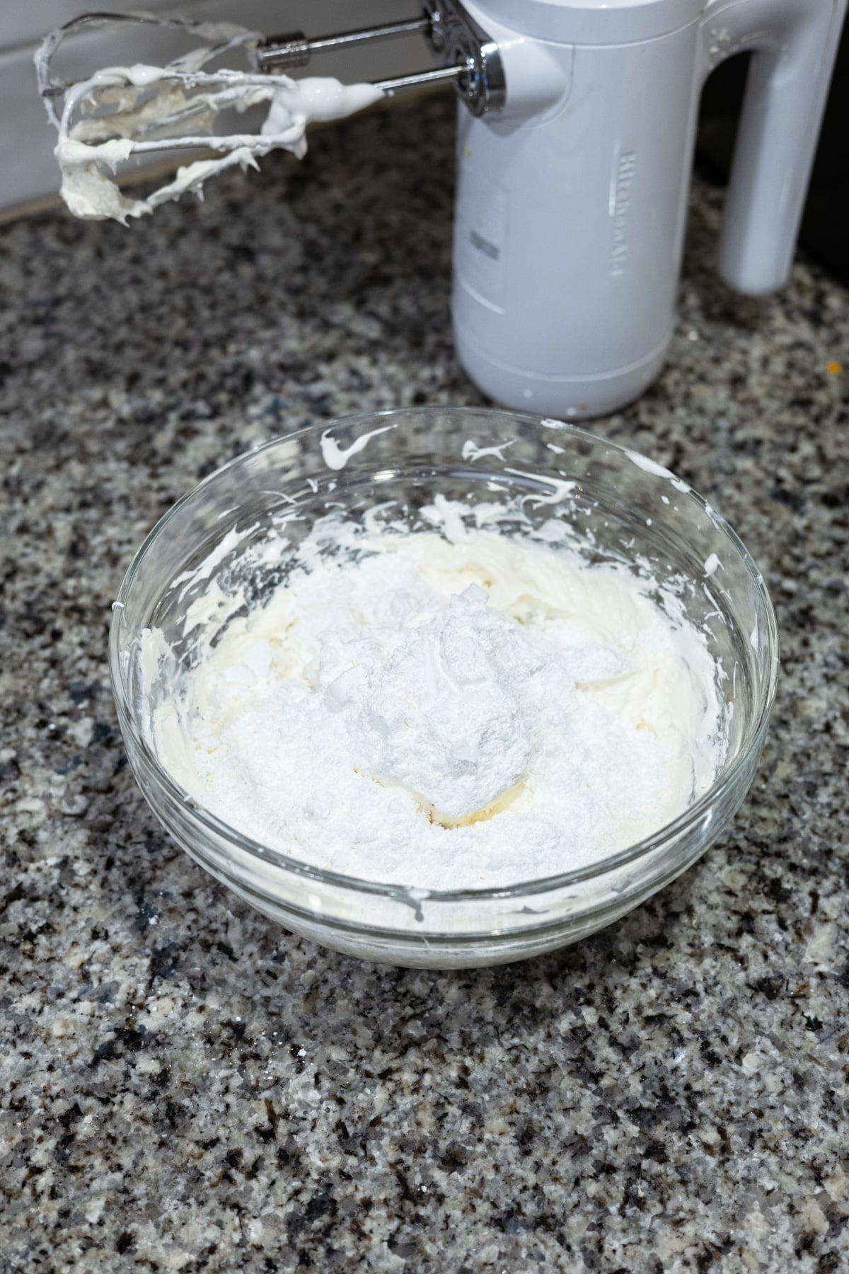 A glass bowl on a granite countertop contains a mixture of white whipped cream and powdered sugar. An electric hand mixer with batter on the beaters rests nearby.
