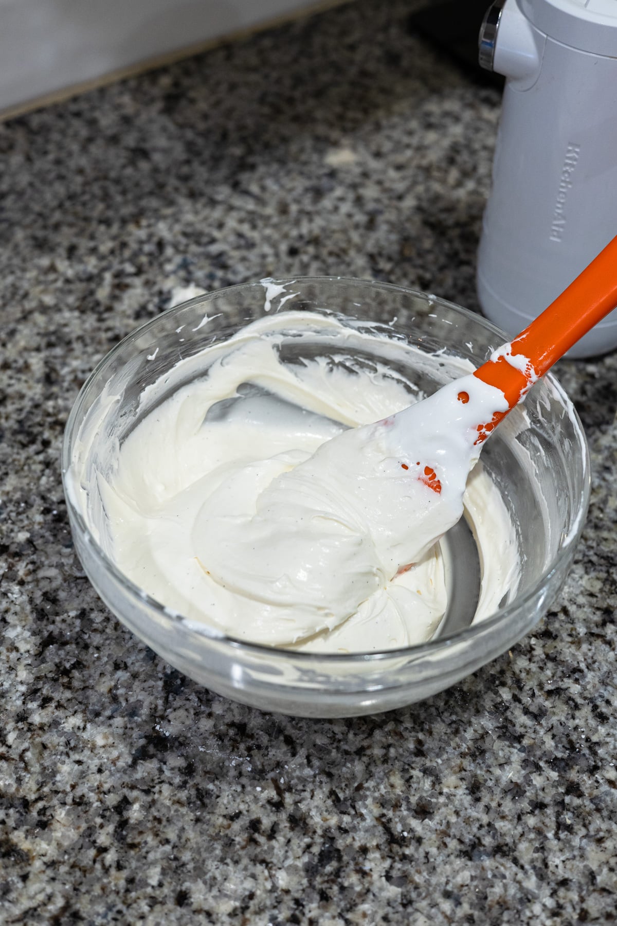 A glass bowl filled with smooth, white whipped mixture sits on a granite countertop. An orange and white spatula rests in the bowl, coated with the mixture. A white kitchen appliance is in the background.