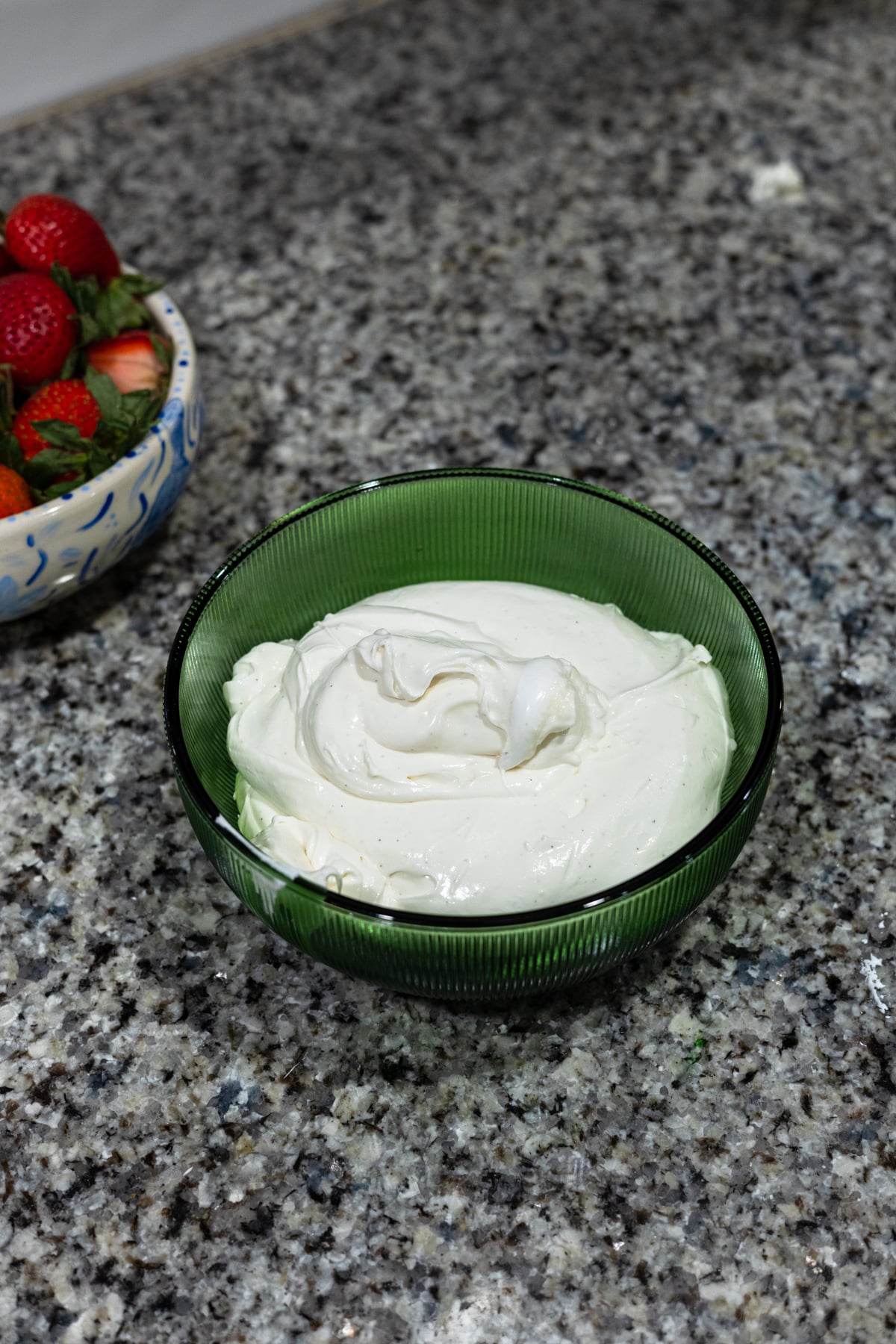 A green glass bowl filled with creamy white dip sits on a granite countertop, with a bowl of fresh strawberries partially visible to the left.