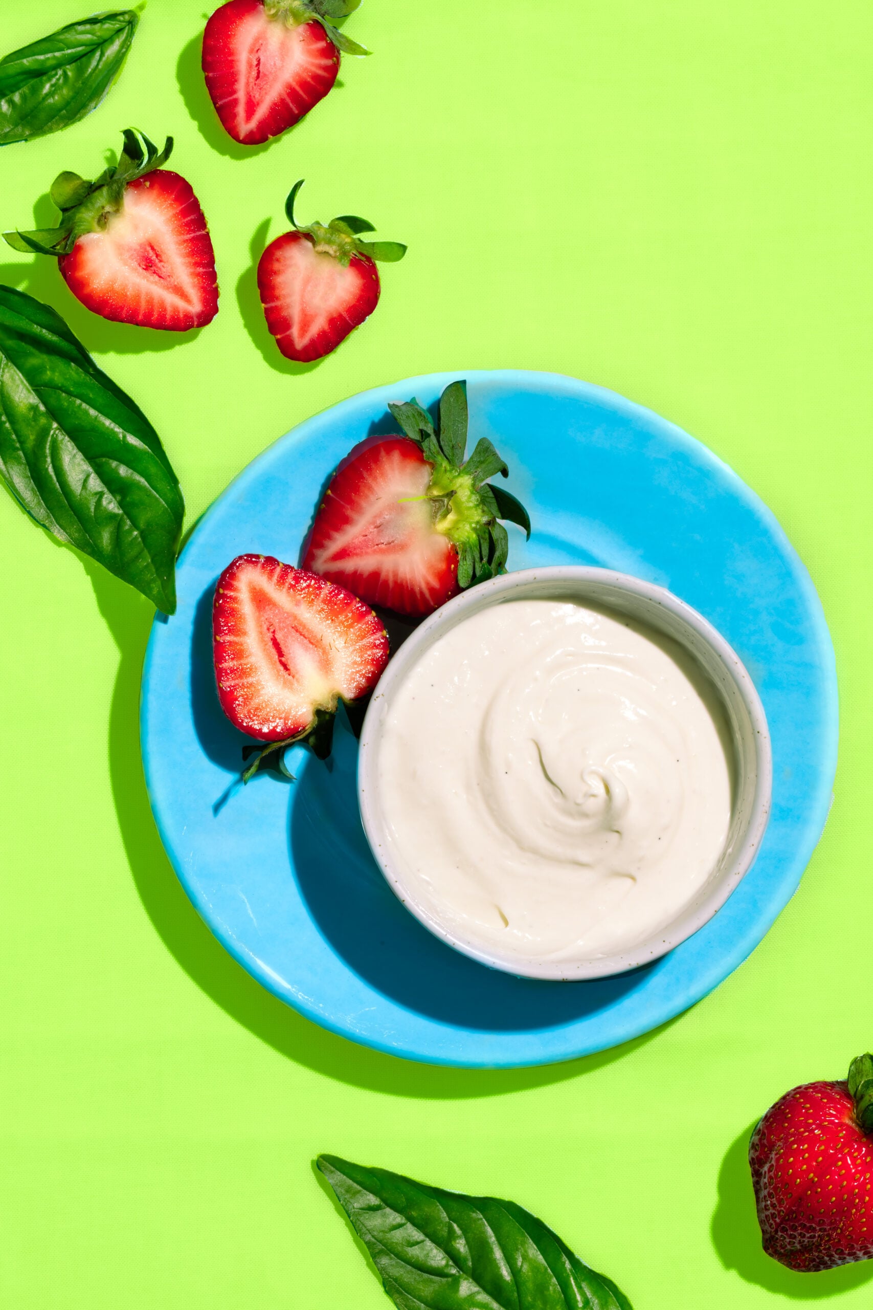 A bowl of creamy white yogurt sits on a blue plate, surrounded by halved and whole strawberries and fresh basil leaves, all arranged on a bright green background.