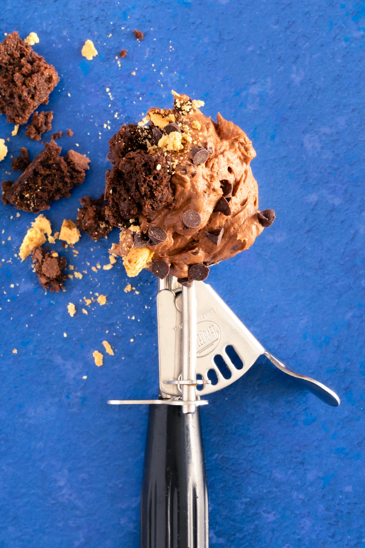 A metal ice cream scoop holds chocolate ice cream with chocolate chips and crumbled cookie pieces, set against a bright blue background.