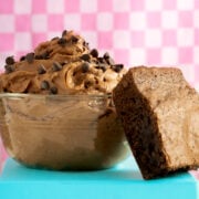 A glass bowl filled with chocolate mousse topped with chocolate chips sits next to a thick, fudgy brownie on a blue surface. Pink checkered background.