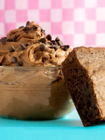 A glass bowl filled with chocolate mousse topped with chocolate chips sits next to a thick, fudgy brownie on a blue surface. Pink checkered background.