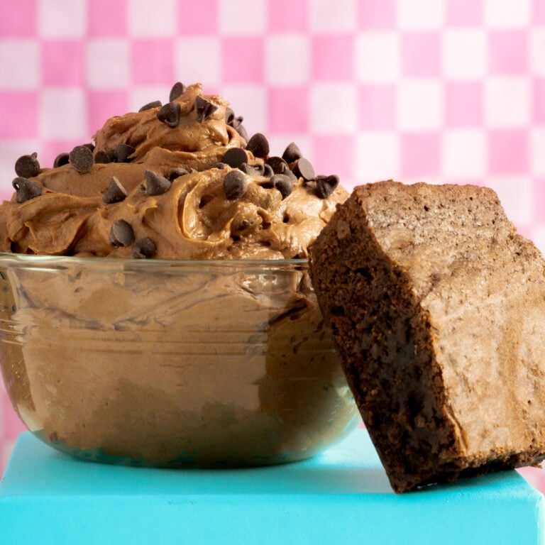 A glass bowl filled with chocolate mousse topped with chocolate chips sits next to a thick, fudgy brownie on a blue surface. Pink checkered background.