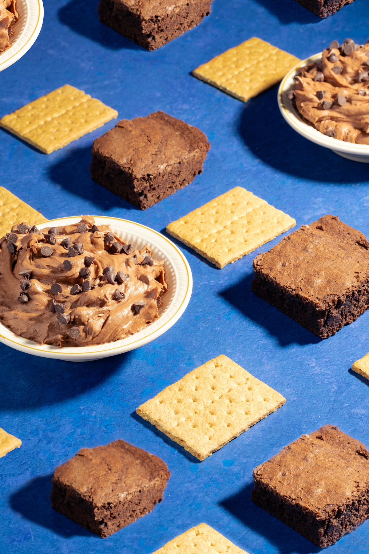Bowls of chocolate mousse with chocolate chips, brownies, and square graham crackers are arranged in a neat pattern on a blue surface.