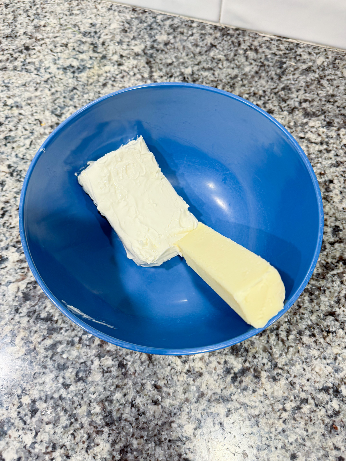 A stick of butter and a block of cream cheese sit side by side in a blue bowl on a speckled countertop.