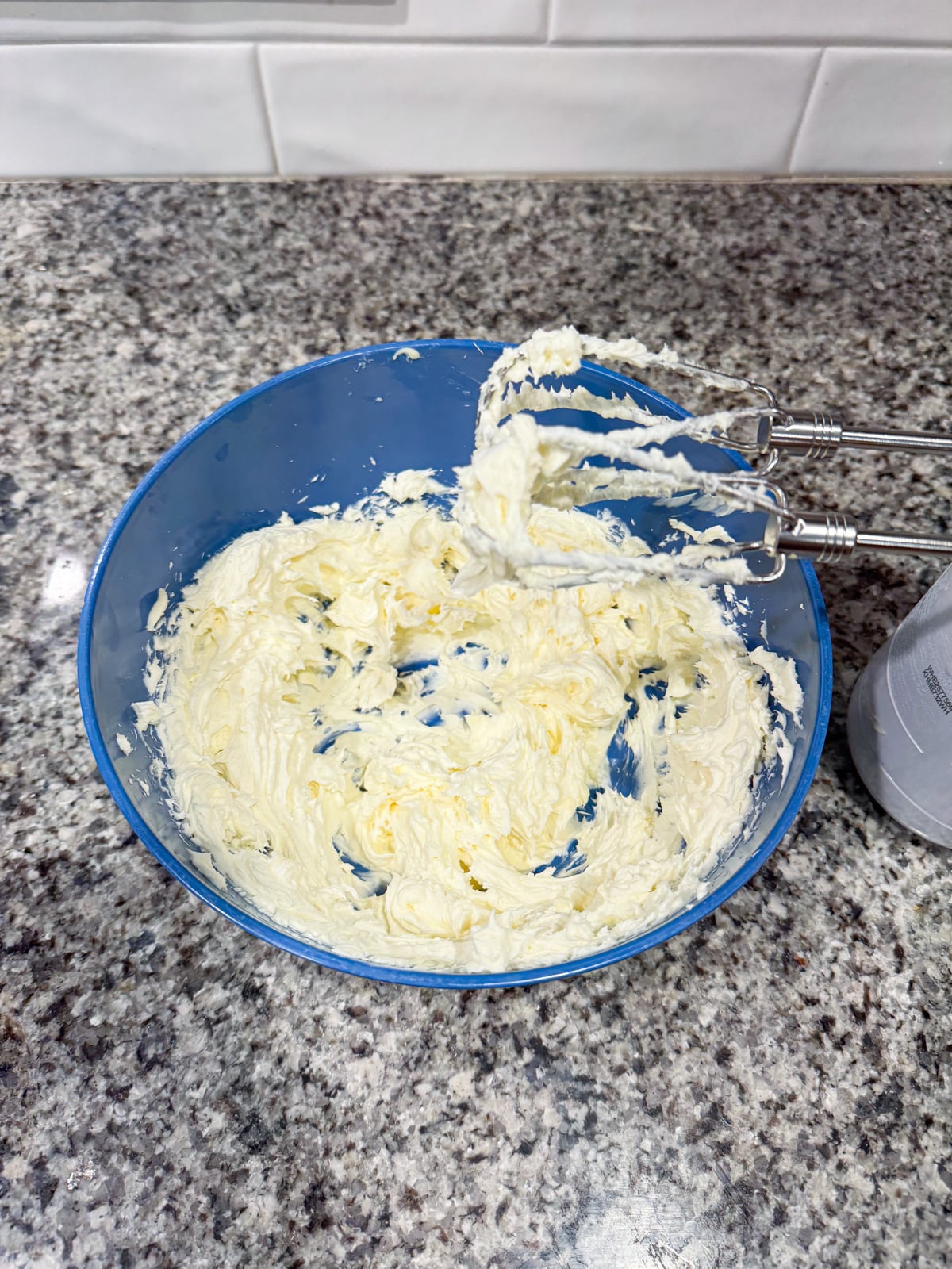 A blue bowl with whipped cream cheese mixture and a hand mixer on a granite countertop. White tiled wall is in the background.