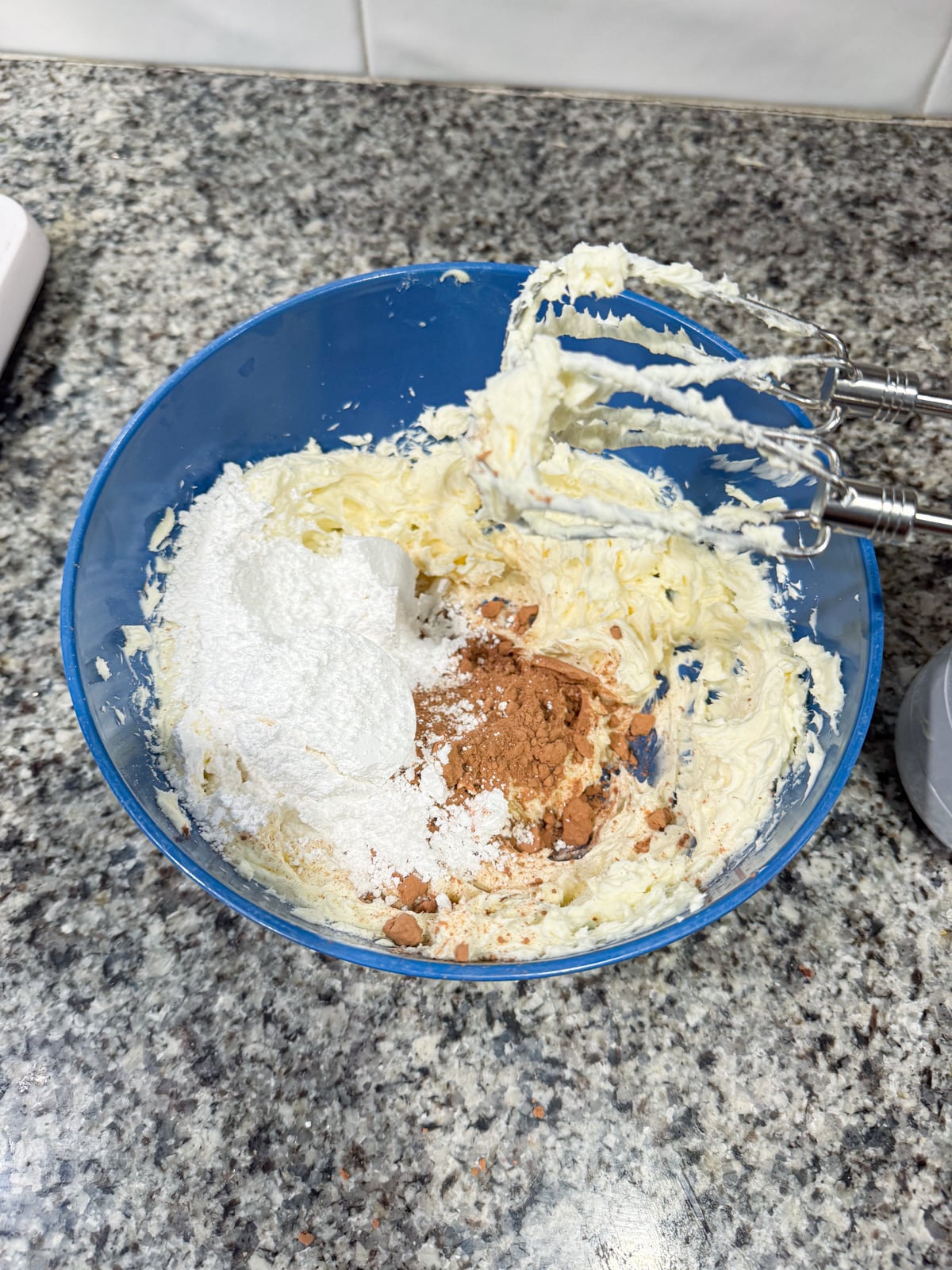 A blue mixing bowl with butter, powdered sugar, and cocoa powder being blended by a hand mixer on a speckled kitchen countertop.