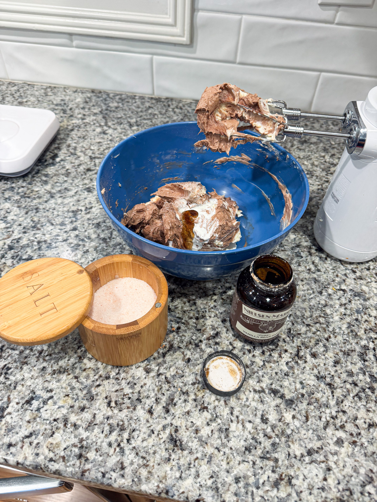 A blue bowl with partially mixed chocolate and white cream is on a granite countertop. Nearby are a salt container, a jar of vanilla extract with its lid off, and a hand mixer with batter on the beaters.