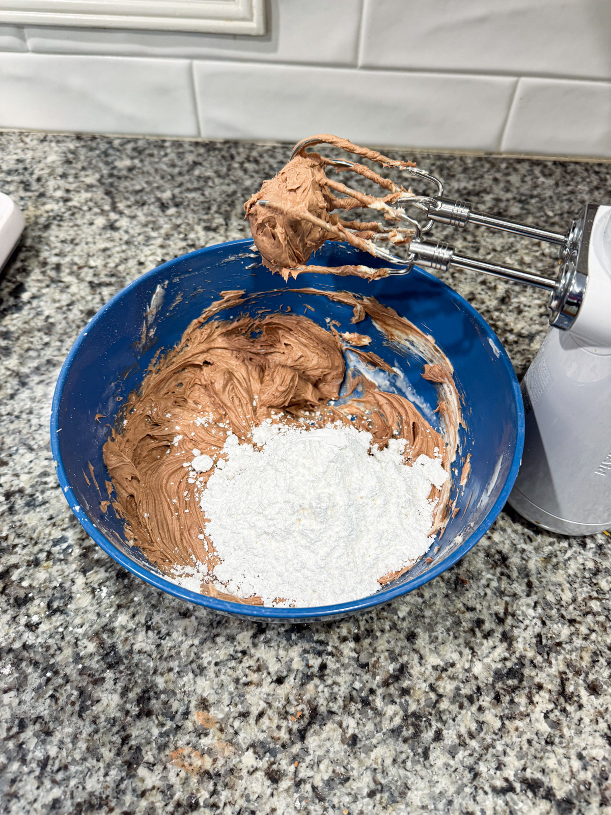 A blue bowl with chocolate batter and a mound of white powdered sugar sits on a granite counter. An electric hand mixer with beaters coated in batter rests on the bowls edge. White tile backsplash is visible in the background.