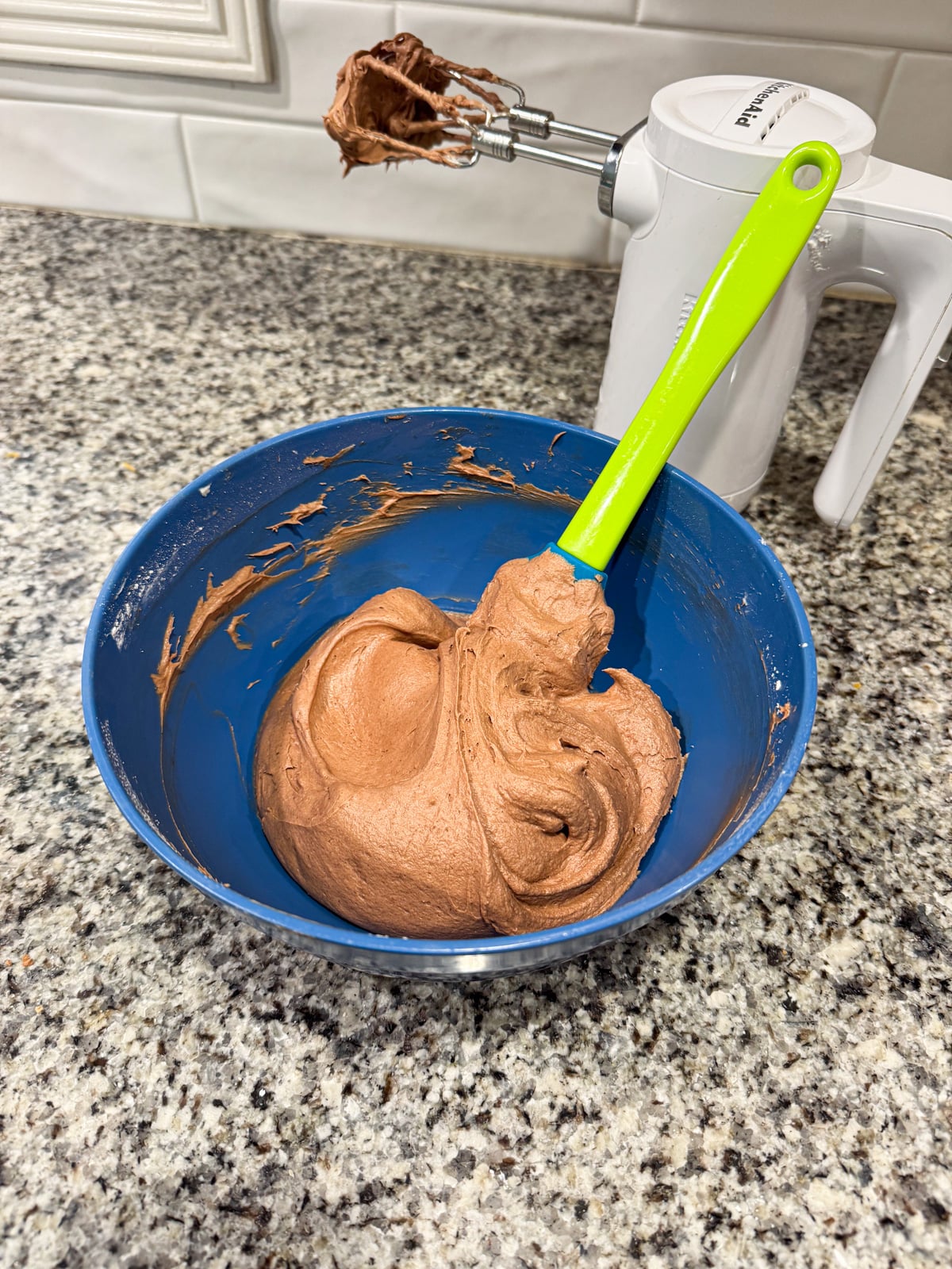 A blue bowl with thick chocolate batter and a green spatula on a granite countertop. An electric hand mixer with chocolate residue is in the background.