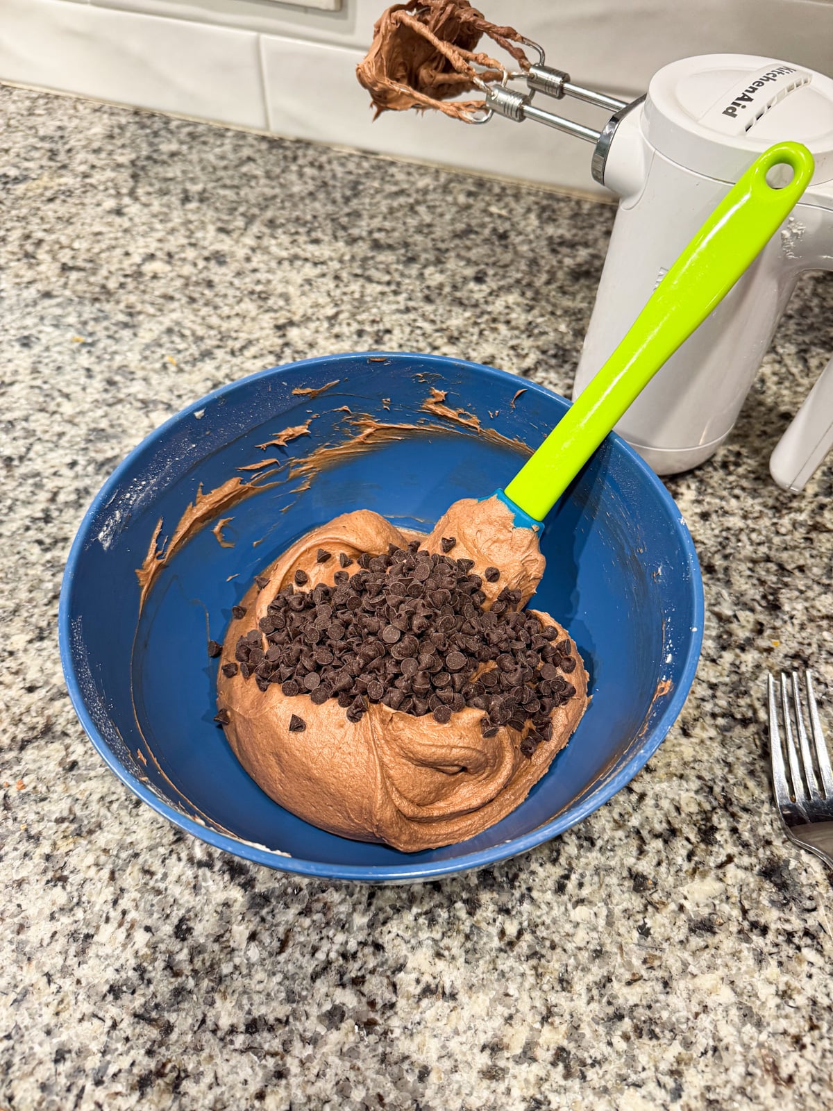 A blue bowl with chocolate cake batter topped with chocolate chips sits on a granite countertop. A green spatula rests in the bowl. An electric mixer with chocolate on the beater and a fork are nearby.