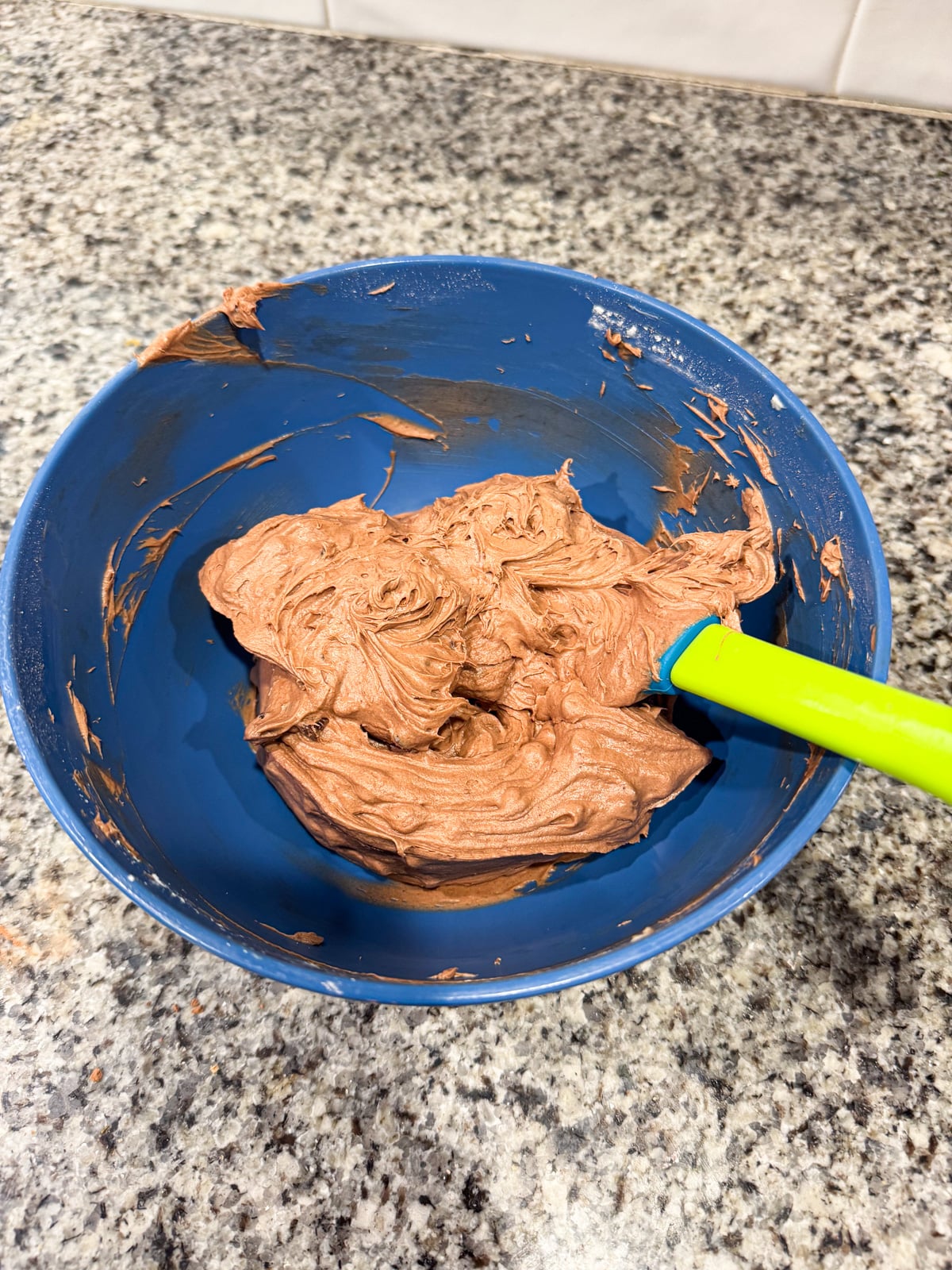 A blue bowl with chocolate cake batter and a green spatula sits on a speckled kitchen countertop.
