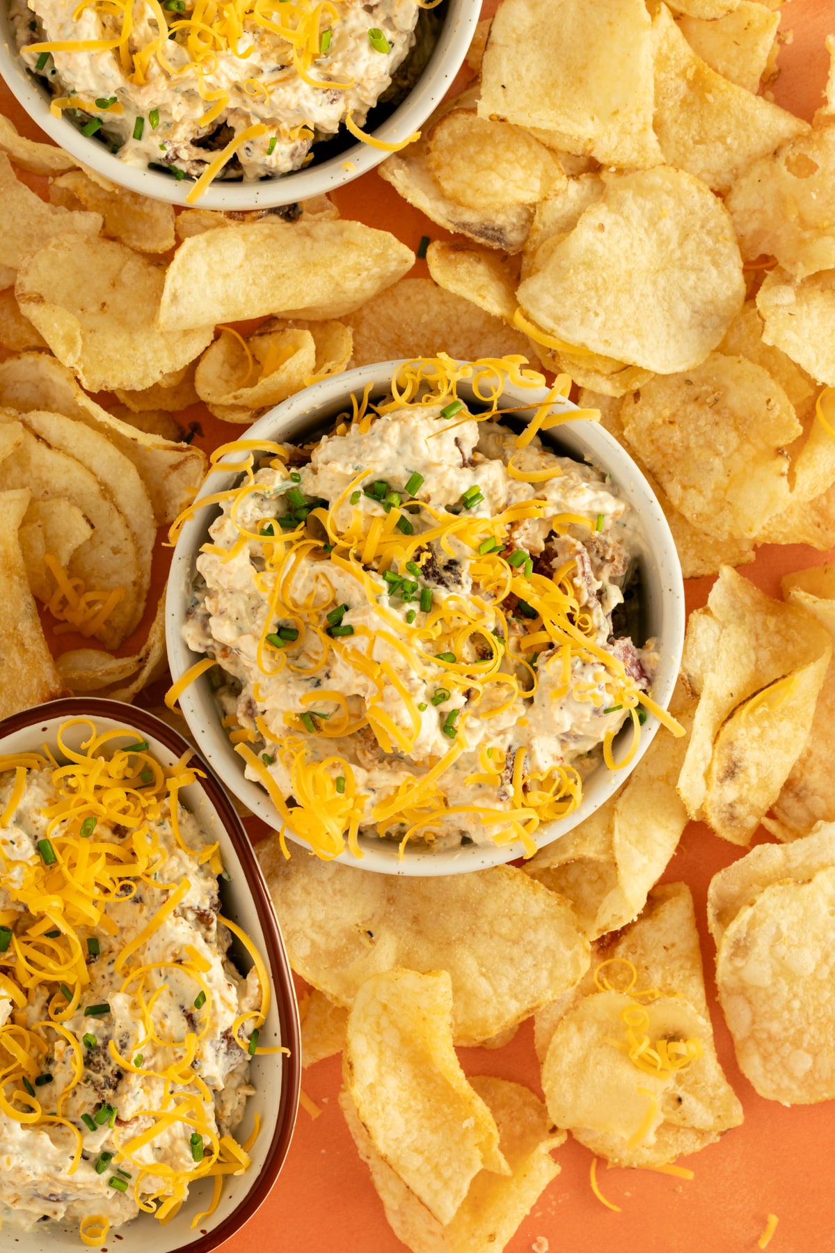 Bowls of creamy dip topped with shredded cheese and chives are surrounded by scattered potato chips on an orange surface.