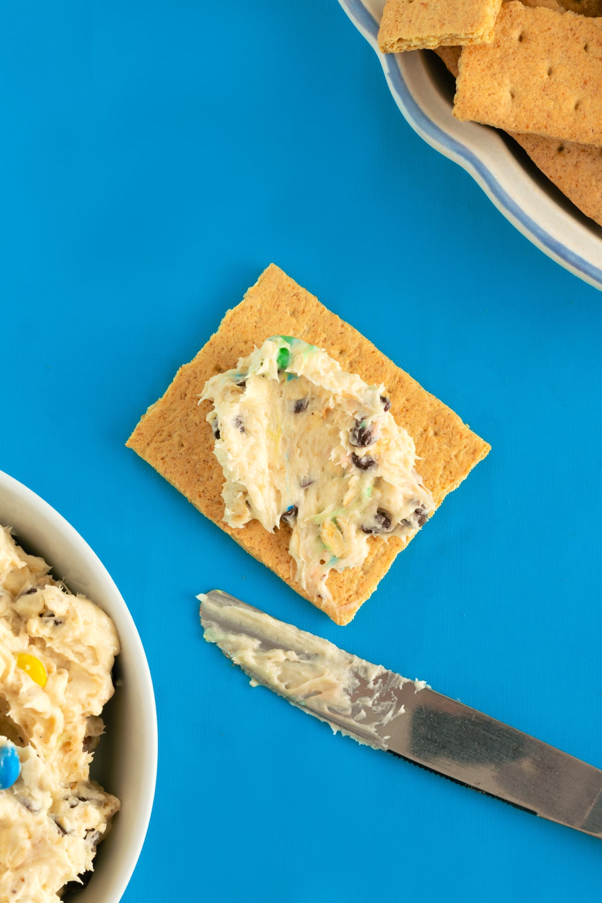 A square graham cracker topped with creamy dip containing chocolate chips and colorful candy, next to a knife with dip on it. A bowl of dip and a dish of more graham crackers are also visible on a blue background.
