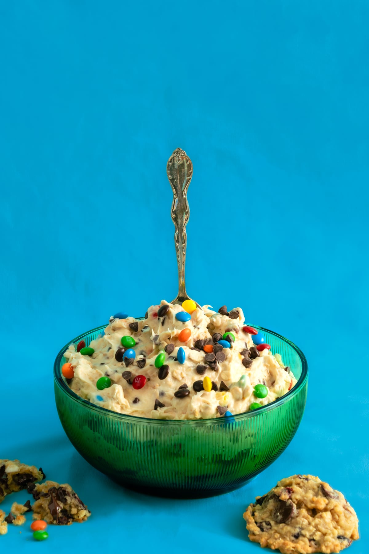 A green glass bowl filled with cookie dough topped with colorful candy pieces, chocolate chips, and a decorative spoon. Broken cookies are placed around the bowl, all set against a bright blue background.