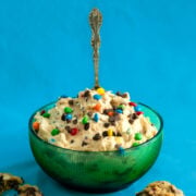 A green glass bowl filled with creamy dessert topped with colorful candy-coated chocolates, with a silver spoon standing upright in the center, against a bright blue background.