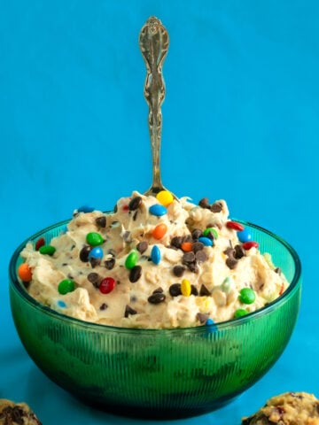 A green glass bowl filled with creamy dessert topped with colorful candy-coated chocolates, with a silver spoon standing upright in the center, against a bright blue background.