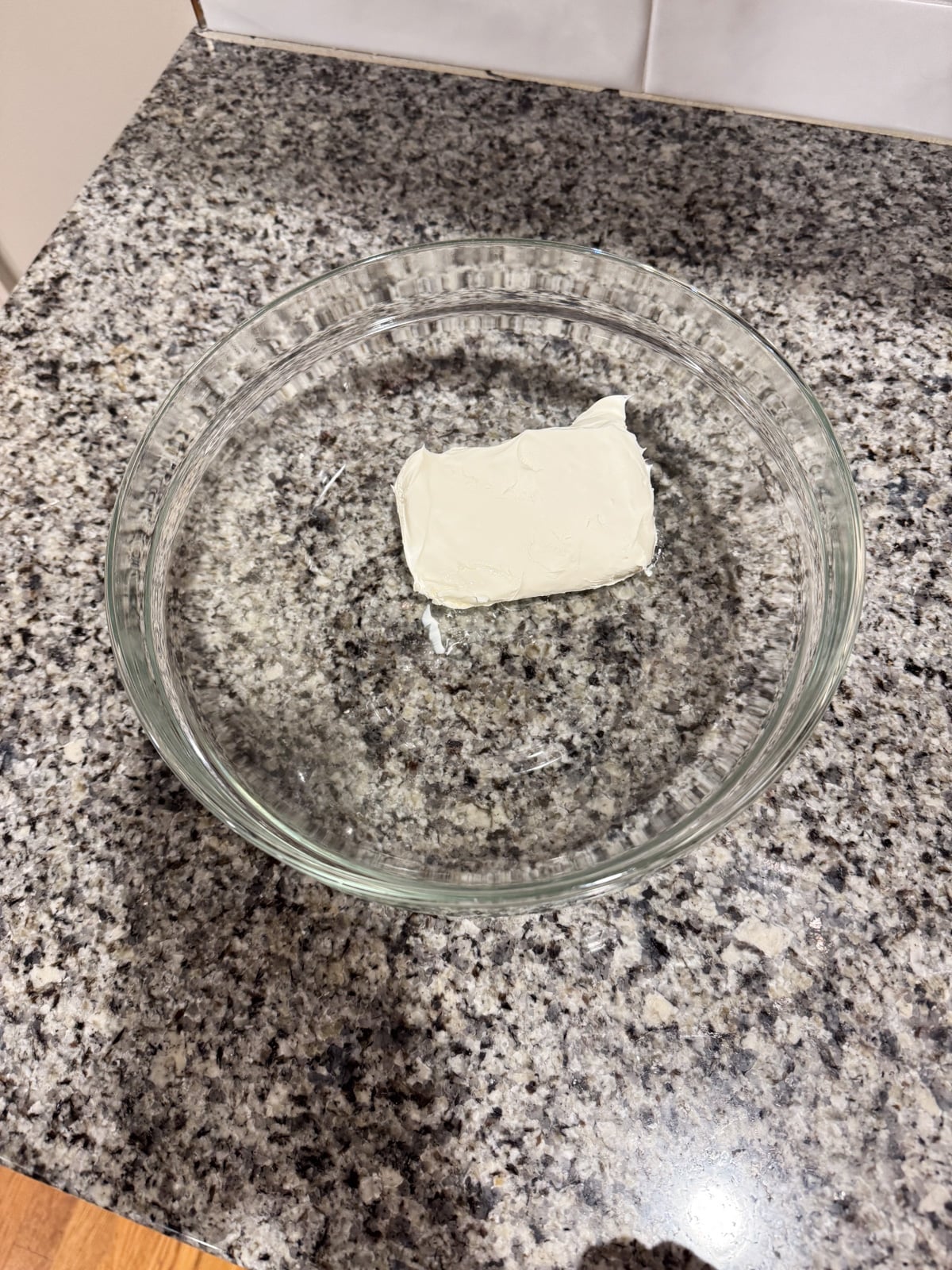 A clear glass bowl sits on a granite countertop, containing a small rectangular block of cream cheese in the center.