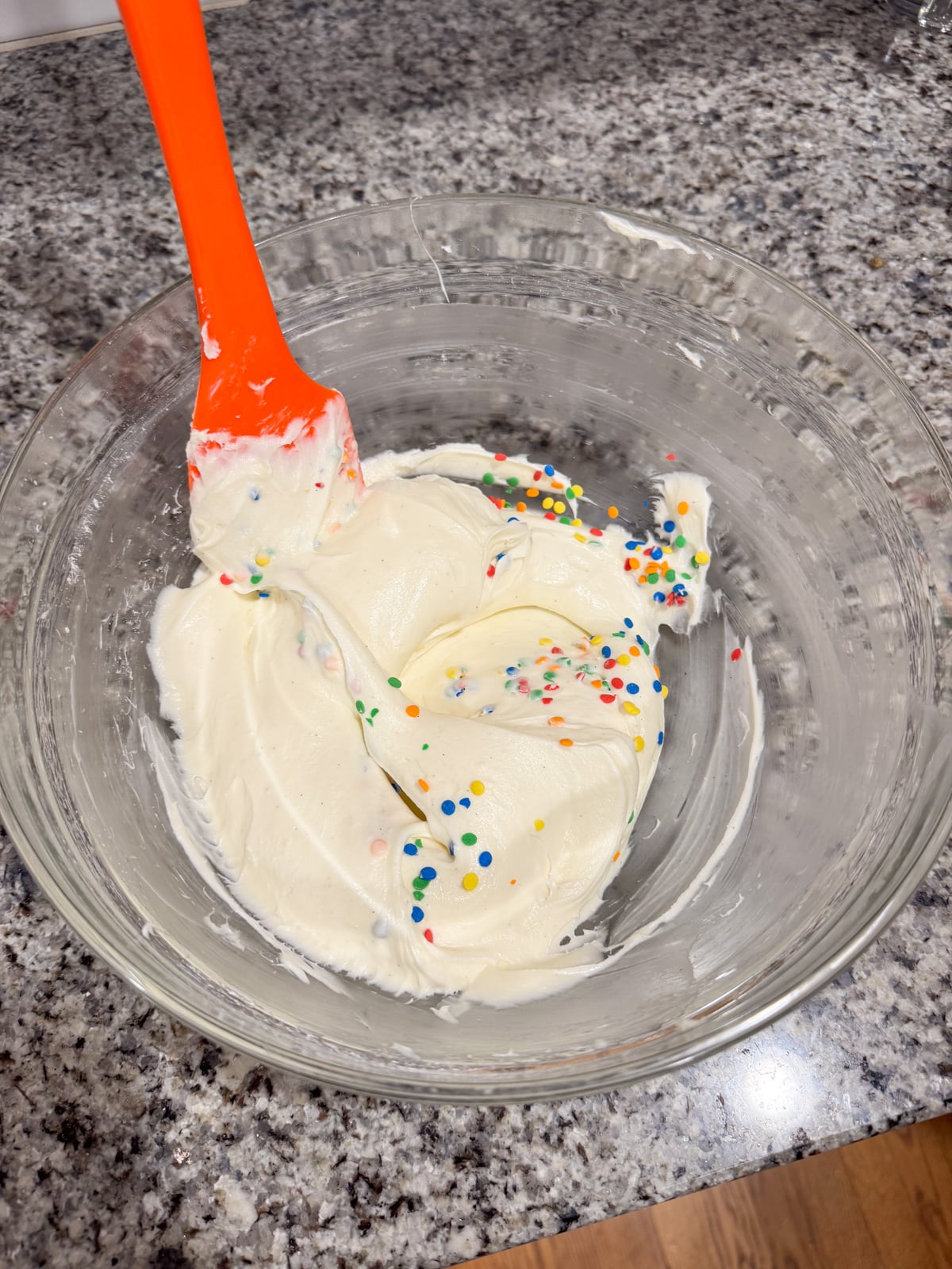 A glass bowl with creamy white frosting, partially mixed with colorful round sprinkles. An orange spatula rests in the bowl on a speckled kitchen countertop.