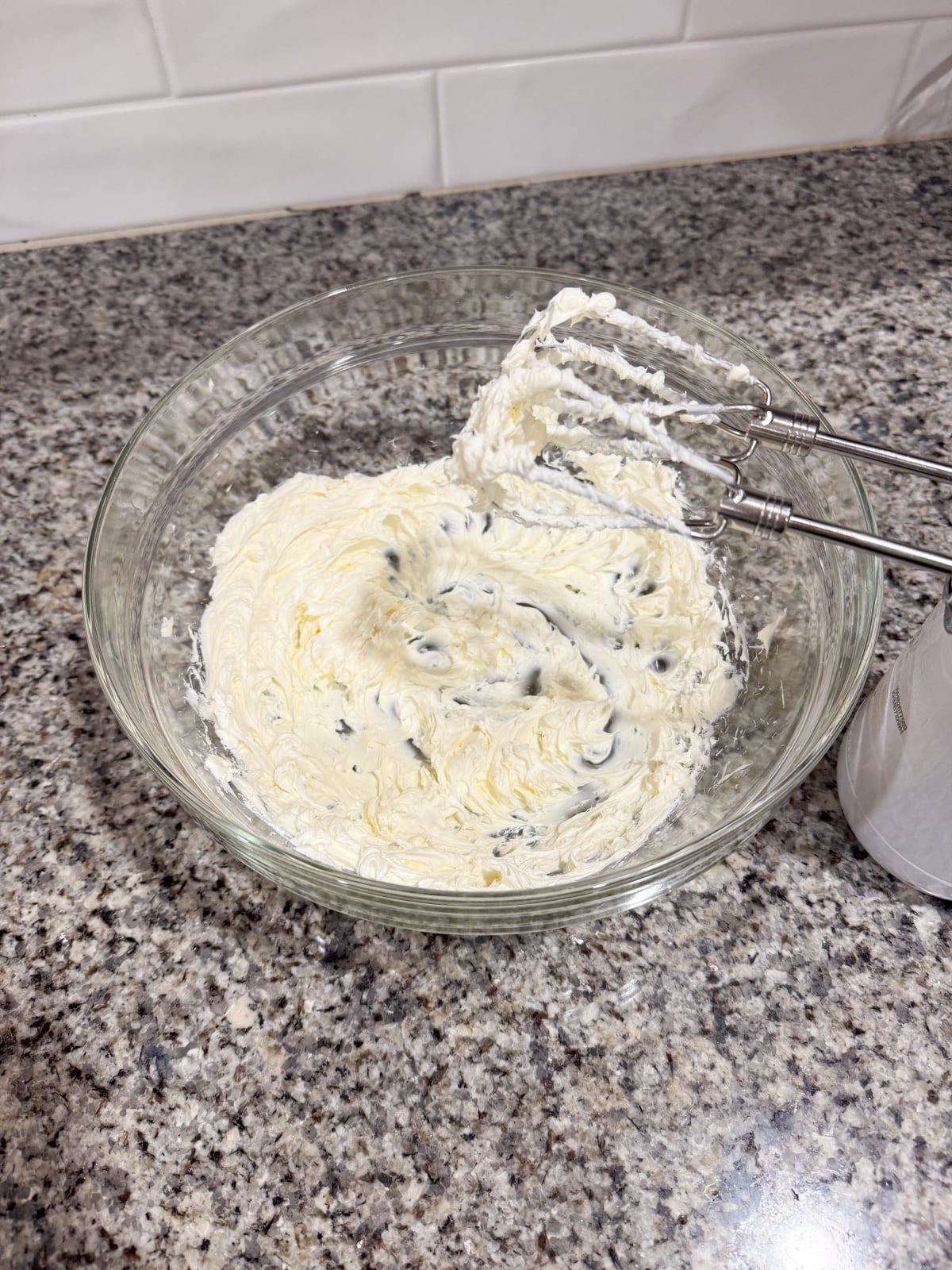 A glass bowl filled with creamy, whipped mixture with chocolate chips sits on a granite countertop. An electric hand mixer with beaters rests in the bowl. White subway tiles are in the background.