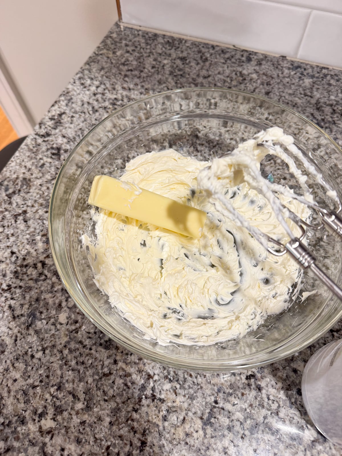 A glass bowl with softened cream cheese and a stick of butter being mixed by an electric hand mixer on a granite countertop.