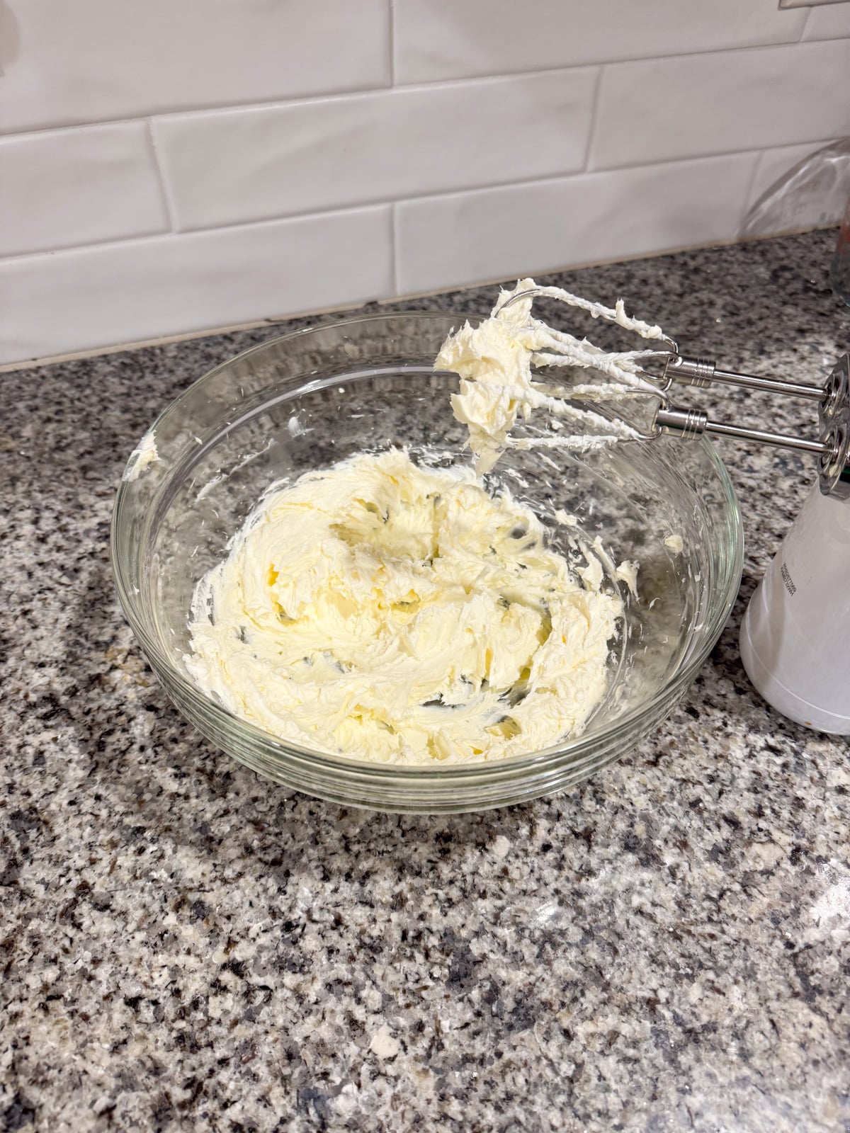 A glass bowl of creamed butter and sugar sits on a granite countertop next to a handheld electric mixer with beaters coated in the mixture. White tiled backsplash is visible in the background.
