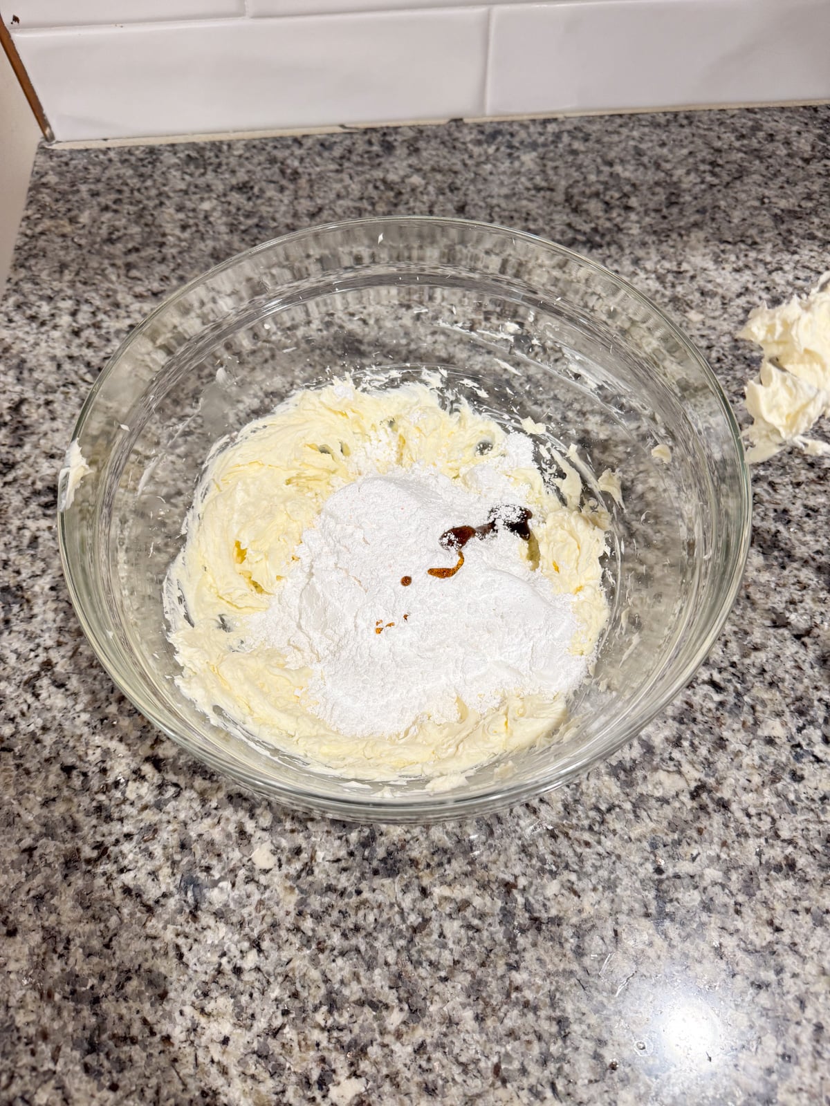 A glass mixing bowl on a granite countertop contains creamed butter, powdered sugar, and a small amount of vanilla extract, ready to be mixed.