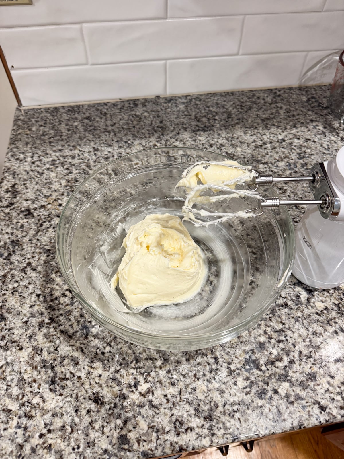 A glass bowl with whipped cream cheese and an electric hand mixer resting on the side sits on a speckled granite countertop in a kitchen with white tile backsplash.