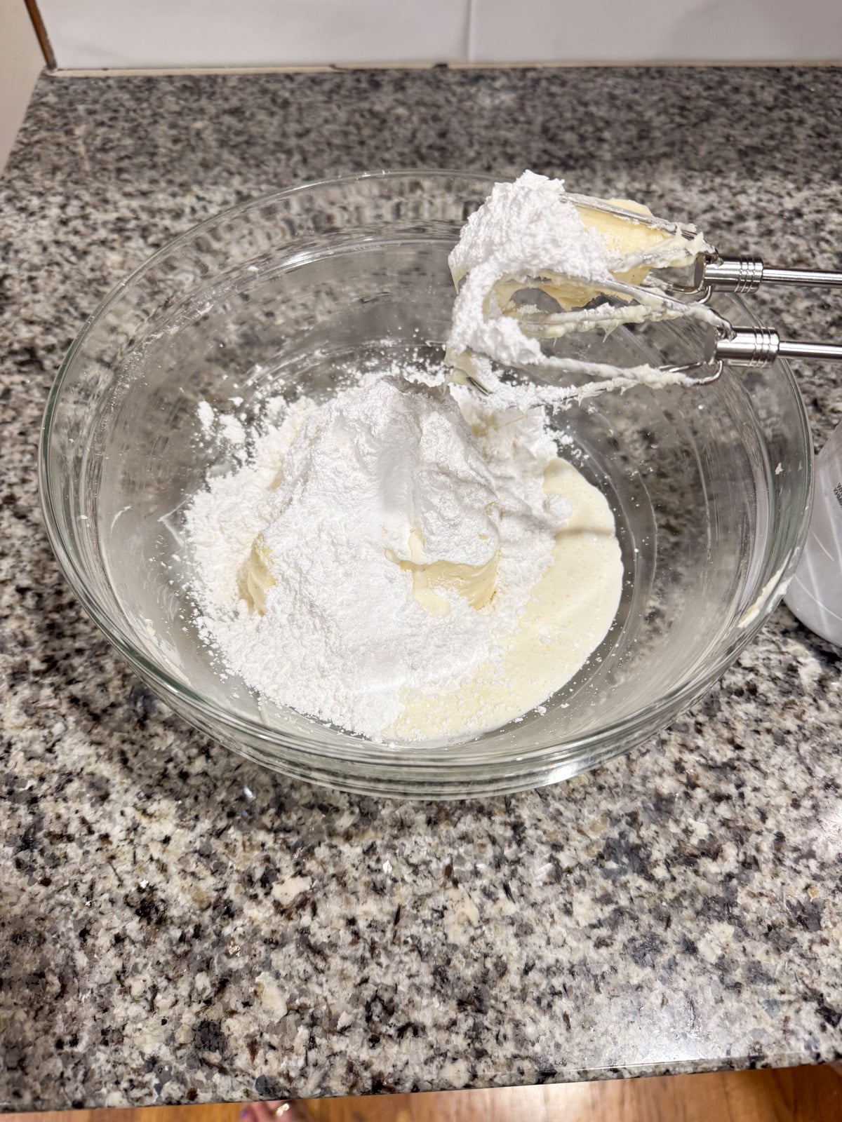A glass bowl on a granite countertop contains powdered sugar, butter, and cream, with an electric hand mixer partially submerged in the mixture, ready for mixing.