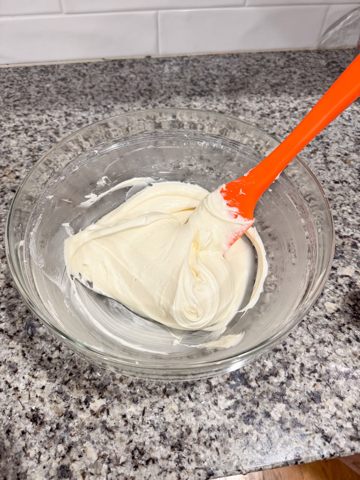 A glass bowl with creamy white frosting being mixed by an orange spatula, sitting on a speckled kitchen countertop with white tile backsplash in the background.