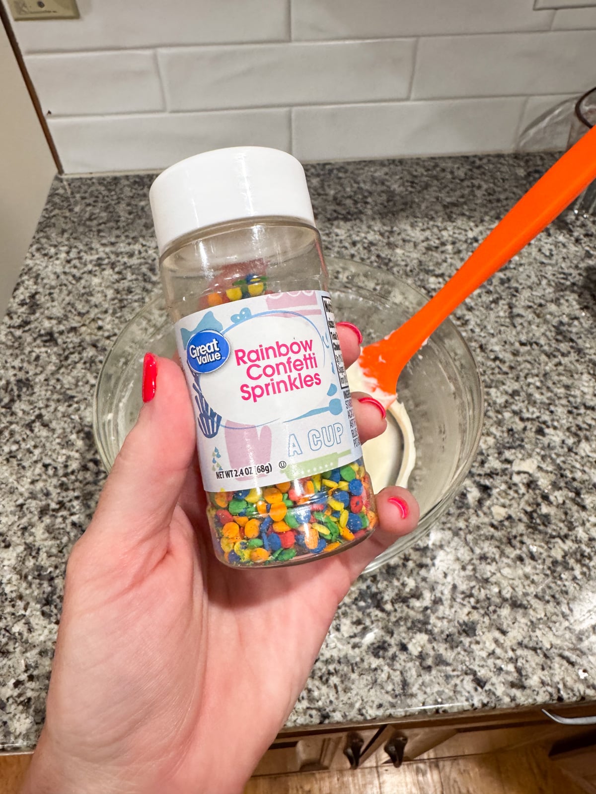 A hand holds a bottle of Great Value Rainbow Confetti Sprinkles over a counter next to a glass bowl of white batter being mixed with an orange spatula.