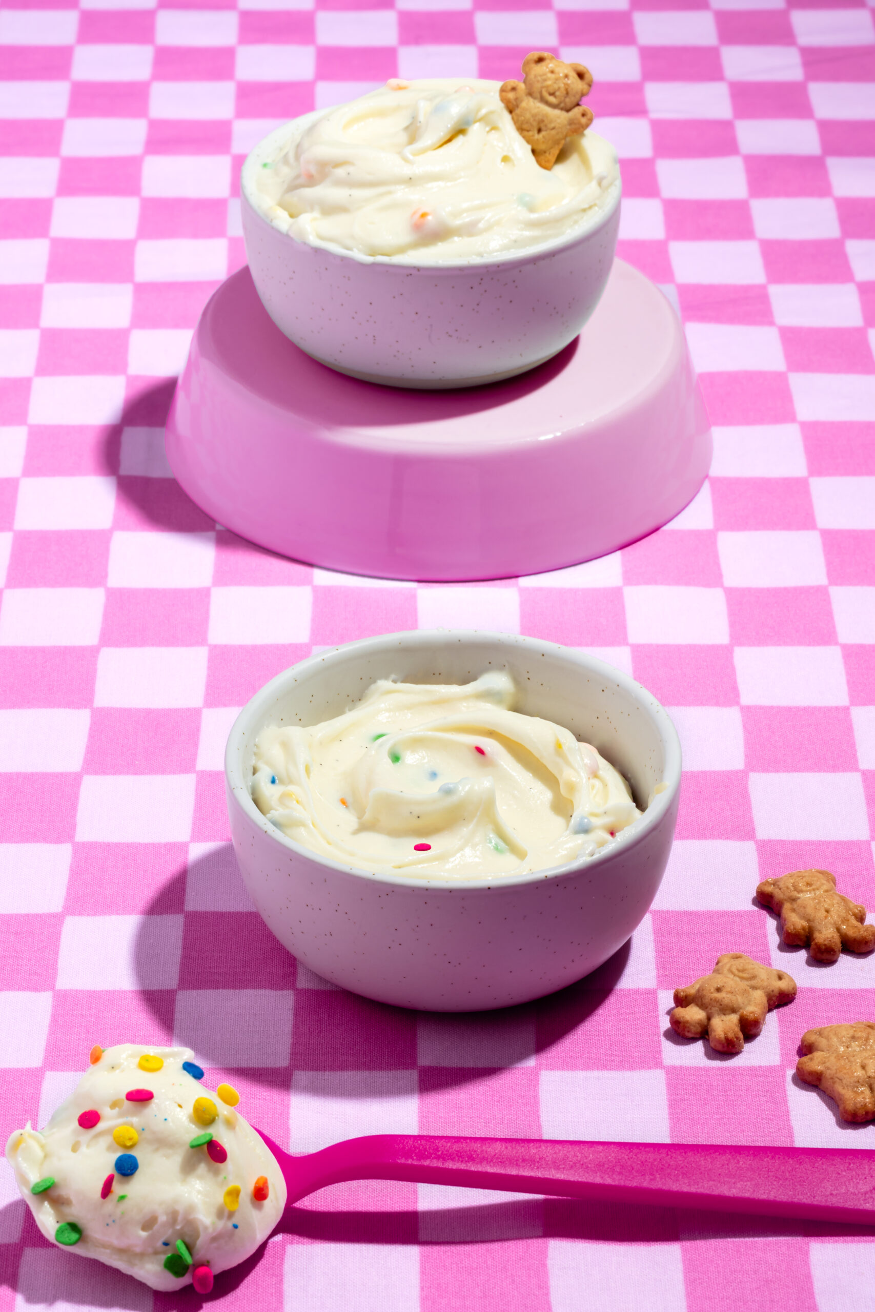 Two bowls of creamy white dessert with sprinkles and teddy bear cookies sit on a pink checkered tablecloth. A pink spoon with a scoop of the dessert is in the foreground. Extra bear cookies are scattered nearby.