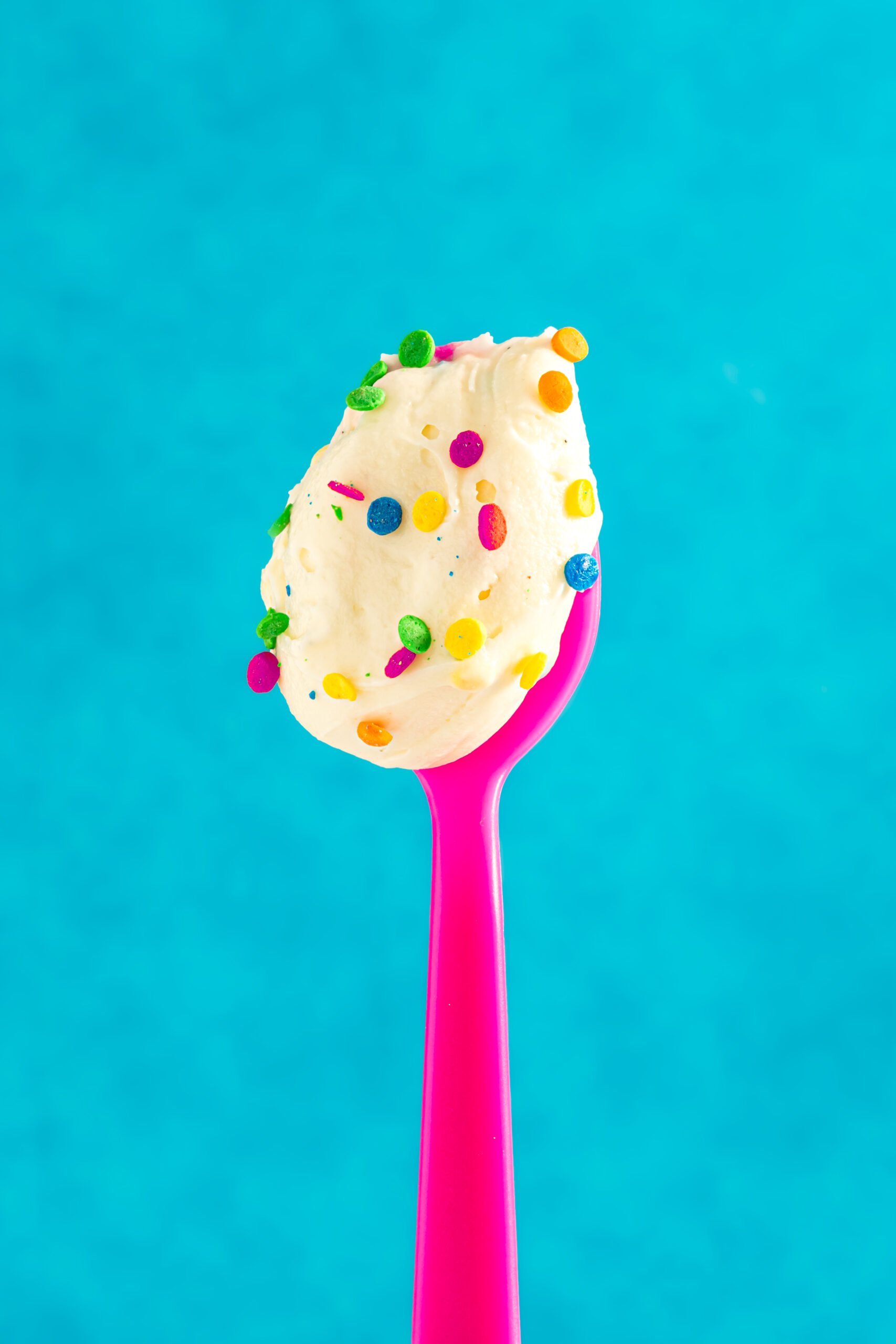 A bright pink spoon holding a scoop of creamy white frosting topped with colorful round sprinkles, set against a vibrant blue background.