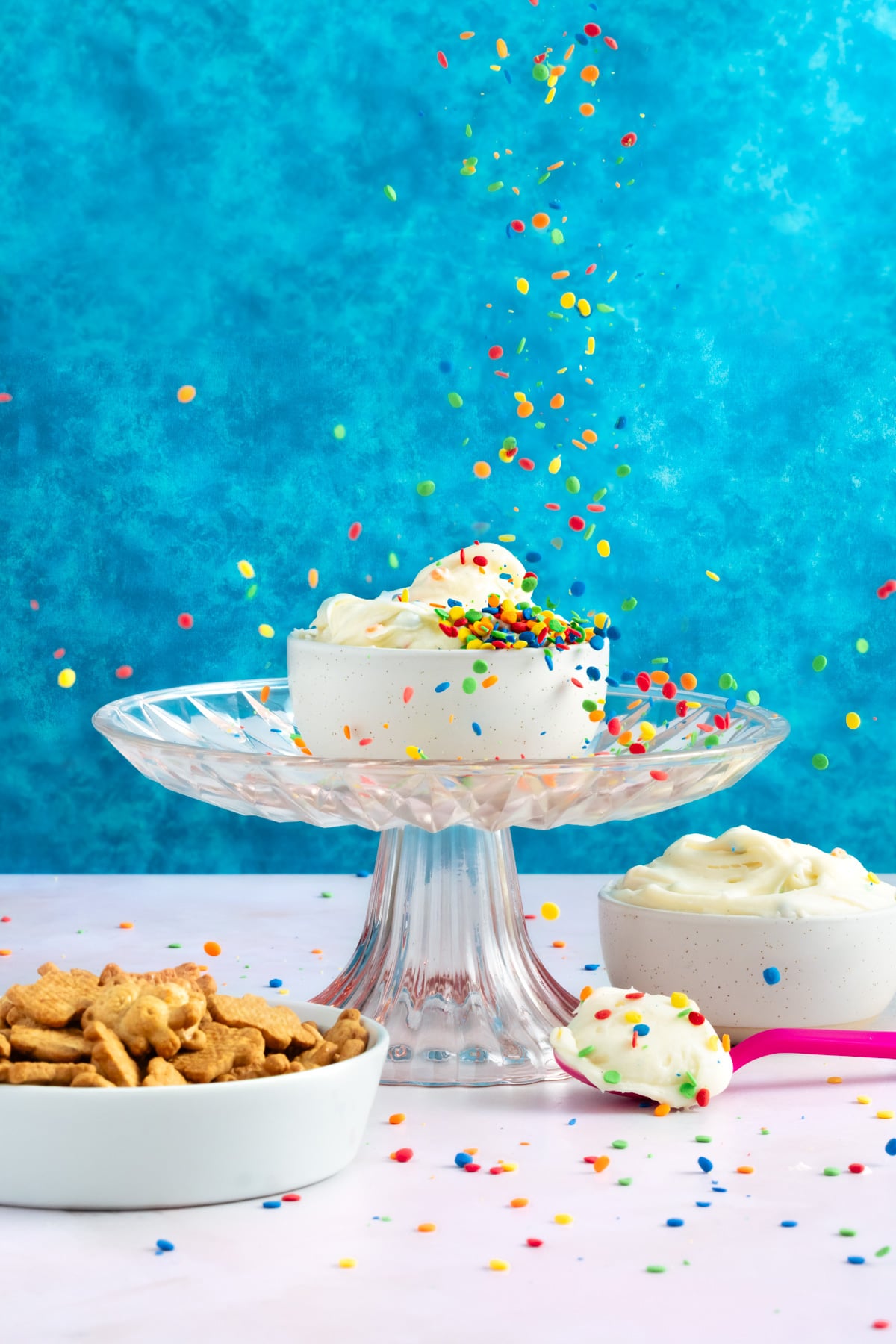 A small cake with white frosting and colorful sprinkles sits on a glass stand, with sprinkles falling onto it. Nearby are a bowl of icing, a spoonful of icing, and a bowl of cereal, all on a pink surface against a blue background.