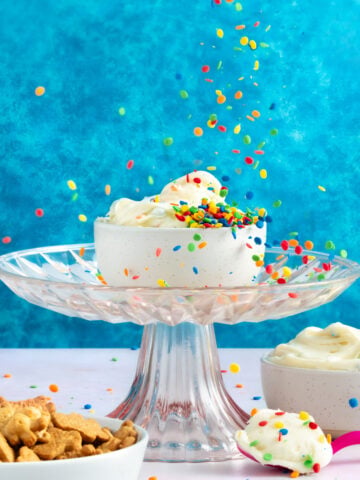 A white bowl of whipped cream with colorful sprinkles sits on a glass cake stand against a bright blue background. Sprinkles fall from above. Nearby are a bowl of whipped cream and a bowl of cereal pieces.