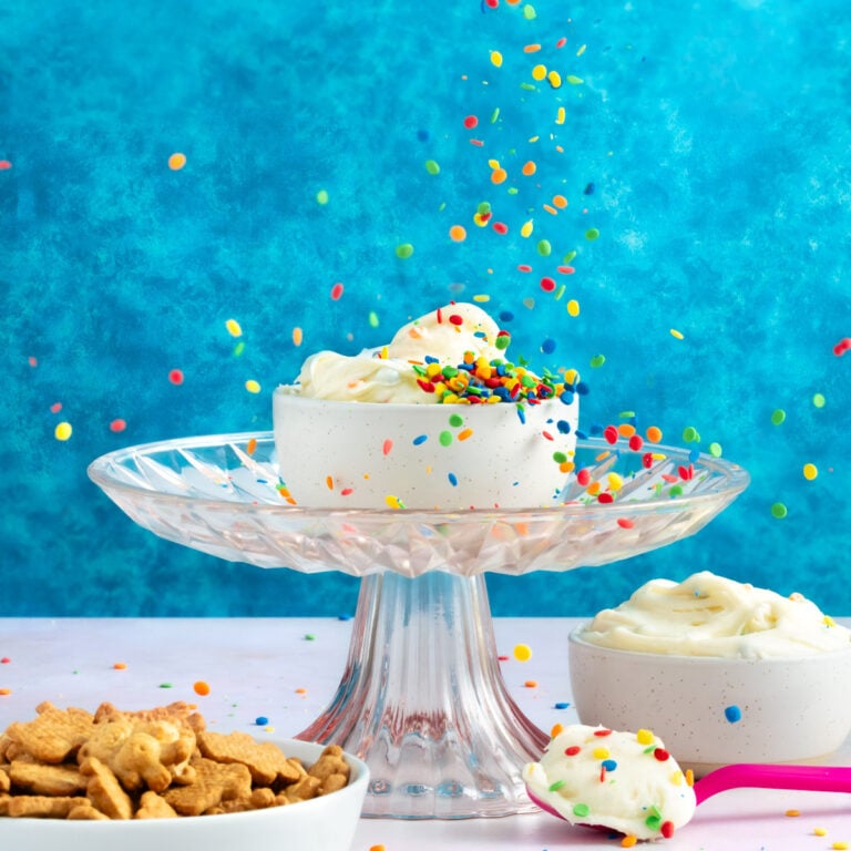 A white bowl of whipped cream with colorful sprinkles sits on a glass cake stand against a bright blue background. Sprinkles fall from above. Nearby are a bowl of whipped cream and a bowl of cereal pieces.